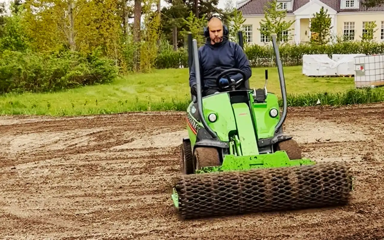 En kompaktlastare sprider toppdress över en gräsmatta. Tunga maskiner och stålskopor ökar risken för markpackning jämfört med manuell utjämning.